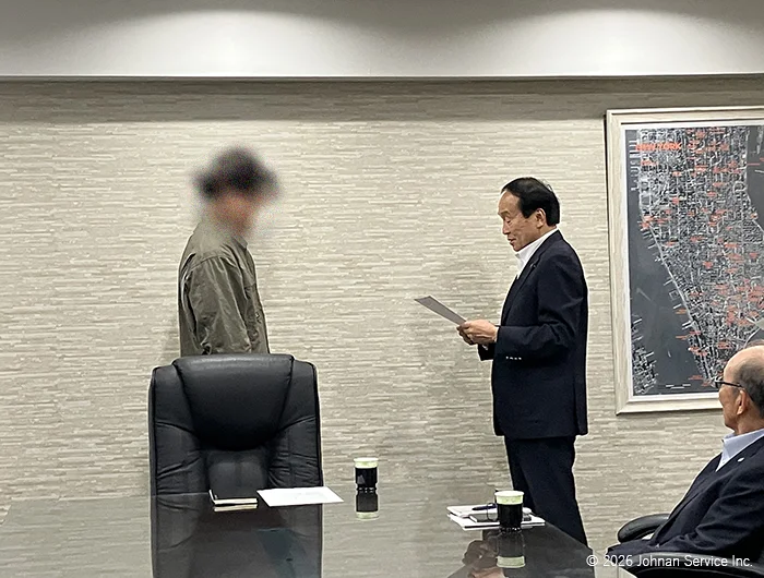 会議室で辞令交付書を手にしたスーツ姿の社長が、立っている男性社員に向かって読み上げる様子。