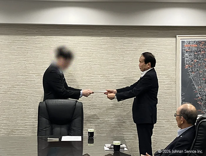 会議室でスーツ姿の社長が、立っている男性社員へ辞令交付書を手渡している様子。