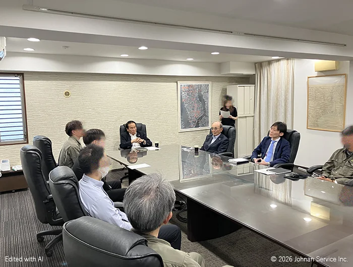会議室のテーブルを囲み、正面の取締役が話し複数の出席者が耳を傾けている様子。