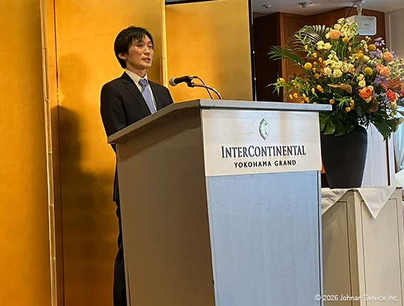 演台に立ち挨拶を行う取締役。背後には金屏風と花が飾られている。