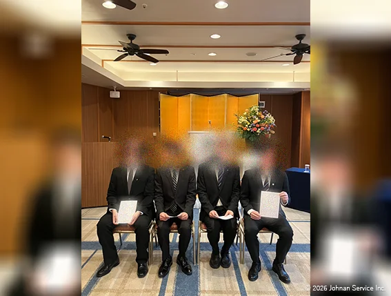 スーツ姿の新入社員4名が椅子に並び、辞令を手にして写る様子。背後には金屏風と演台が設置されている。