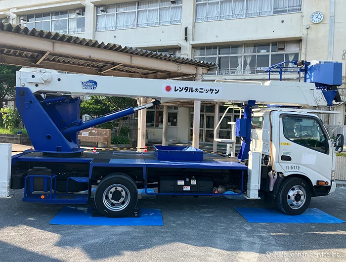 校舎前に停められた白と青の高所作業車。前後のタイヤの下にブルシートを敷いている。