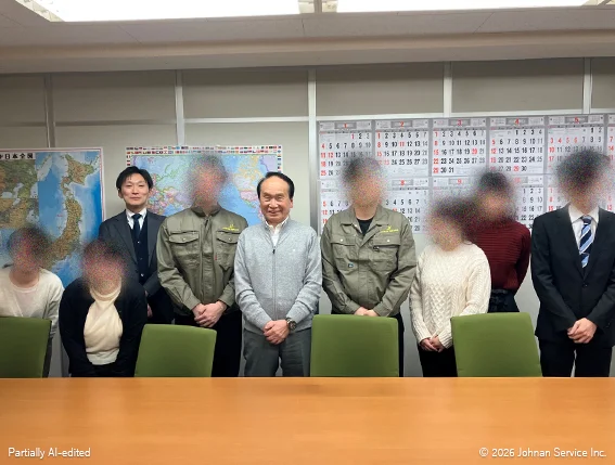 9人が横一列に並んで記念撮影している様子。背後には地図やカレンダーが貼られ、手前に会議テーブルがある。