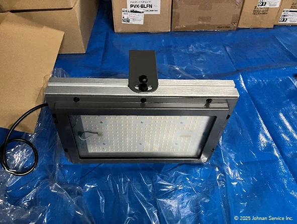 青いブルーシート上に置かれた長方形のLED照明器具。金属フレームとカバーがあり、LEDアレイが見える。上部には黒い取り付けブラケットがあり、一方の側から電源ケーブルが伸びている。