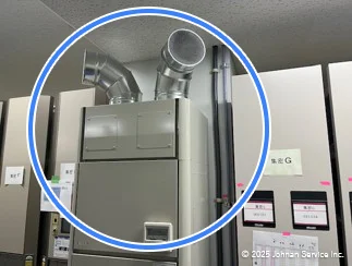 屋内の一角を写した画像で、青い円で強調された中央には、通気ダクトが上部に接続された金属製の除湿器が設置されています。その周囲には、ラベルや書類が貼られた複数の制御パネルが並んでいます。
