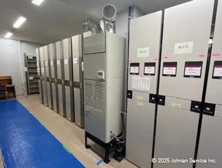 蛍光灯で明るく照らされた室内に、複数の制御パネルが並んでいます。左側の床にはブルーシートが敷かれており、中央には通気ダクトが接続された大型の金属製除湿器が設置されています。