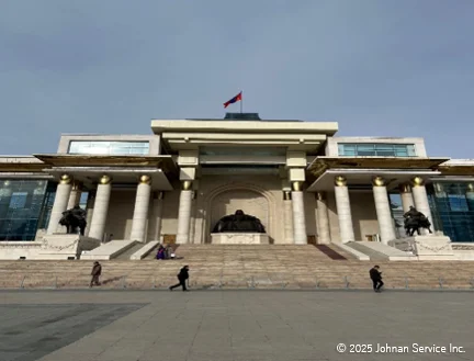 モンゴルの政府宮殿の正面外観。中央に階段があり、上部にはモンゴル国旗が掲げられた大きな建物。入り口前には騎馬像が左右に配置され、白い柱が並ぶ壮麗な構造。