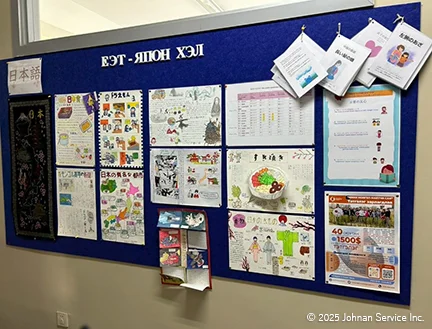 青い掲示板に日本語学習関連の資料やイラストが多数貼られている様子。手書きのポスター、カラフルな絵、印刷物が並び、上部には日本語とモンゴル語の文字が表示されている。