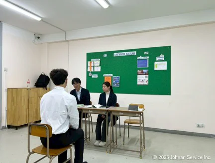 教室内での面接の様子。緑の掲示板を背景に、スーツ姿の面接官2人が机に座り、白シャツの受験者が椅子に座って向かい合っている。