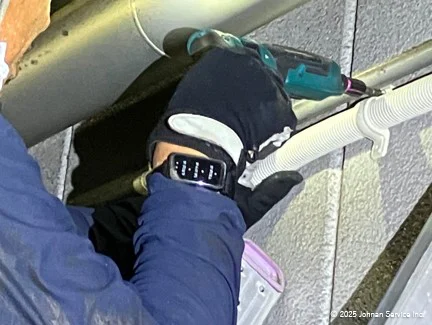 青い作業着と黒い手袋を着用した人物が、右手首に熱中症対策バンドを装着し、電動ドリルでサドルバンドを取り付けている様子。