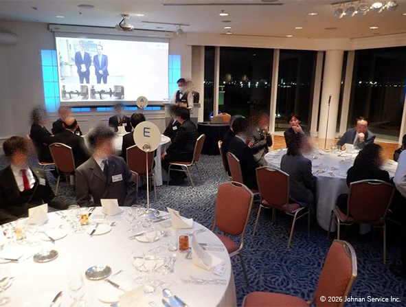 円卓が並ぶ会場で多くの参加者が着席している様子。テーブルにはカトラリーやメニューが整えられ、会場奥には大きな窓から夜景が見える。