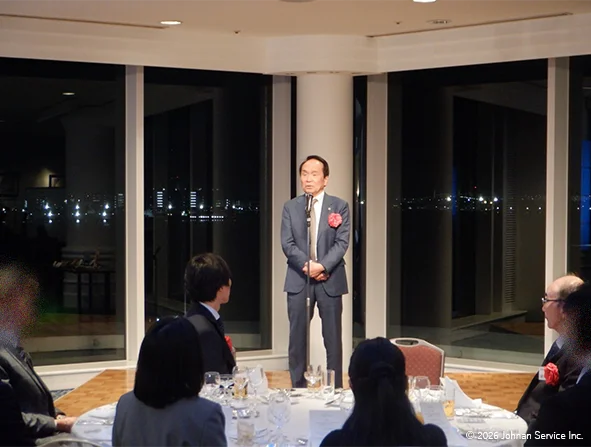 夜景が見える大きな窓の前でスーツ姿の社長がマイクの前に立ち挨拶をしている様子。円卓にはグラスや食器が並び、周囲の参加者が着席して話を聞いている。