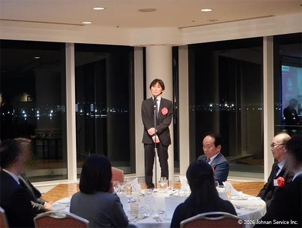 夜景が見える大きな窓の前でスーツ姿の取締役がマイクの前に立ち挨拶をしている様子。