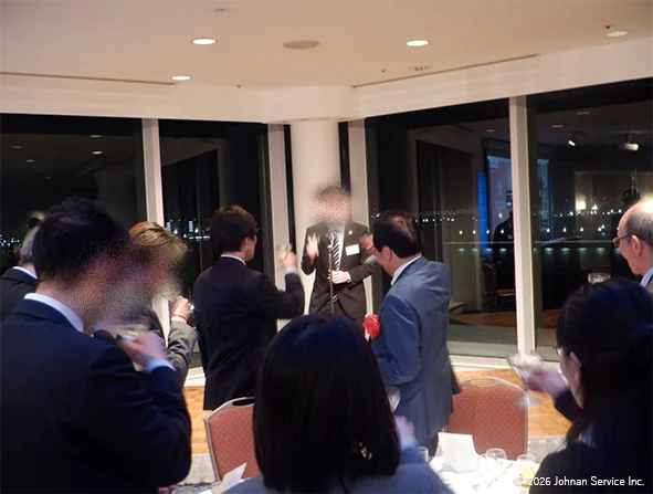 スーツ姿の人物がマイクの前で挨拶をしており、周囲では参加者たちがグラスを持って立ち上がり乾杯の場面を迎えている様子。