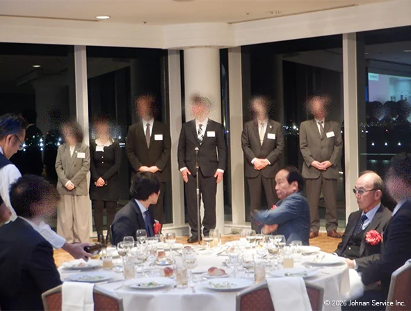 夜景が見える大きな窓の前で数名の参加者が前方に横一列で立ち登壇している様子。
