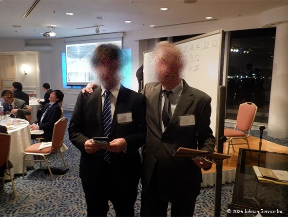 二人のスーツ姿の人物が肩を寄せて立ち、ビンゴゲームの景品を手にしながら楽しそうに記念撮影をしている様子。