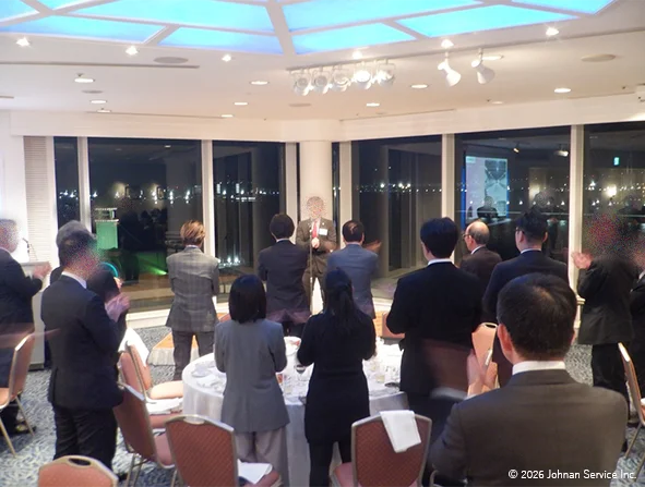 円卓が並ぶ会場で参加者たちが立ち上がり、拍手を送りながら前方のスーツ姿の社員に注目している様子。